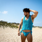 Man wearing a blue crochet tank top and swim briefs on a sandy beach.