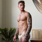 Man with tattoos wearing beige underwear in a room with plants and furniture.